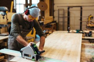 A carpenter using a circular saw on a recycled pallet in a woodworking workshop, wearing safety headphones and goggles.