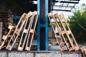 Stacks of wooden pallets leaned against a blue metal structure in an industrial area with a blurred background.