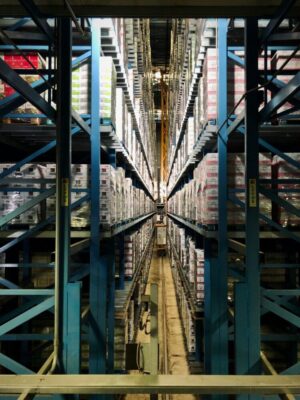 The interior of the large warehouse features high shelves stacked with boxes, emphasizing its role in sustainable supply chains. A central aisle runs through the middle, and at the far end, a crane operates efficiently alongside eco-friendly pallets.