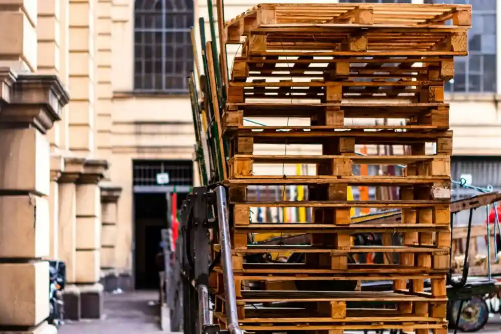 A stack of wooden pallets is loaded onto a cart on a city street near a beige building with large windows.