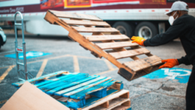 A person wearing gloves and a mask moves a wooden pallet onto a stack of pallets in a parking lot near a truck.