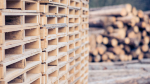 A stack of wooden pallets is shown in the foreground, with blurred logs piled in the background outdoors.