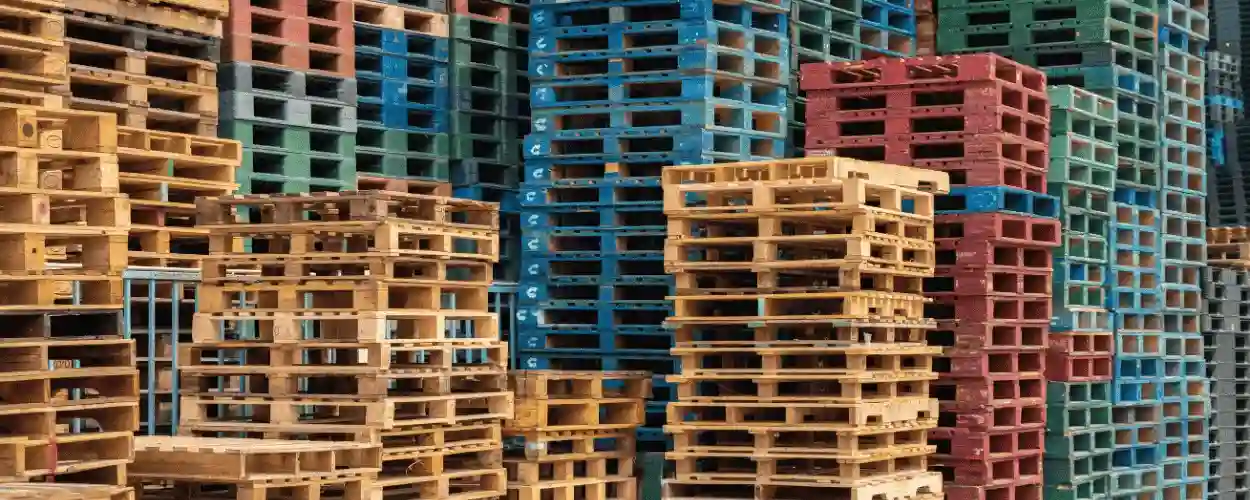 Stacks of wooden and plastic pallets in various colors and sizes are arranged closely together outdoors, showcasing a well-organized pallet supplier’s inventory.