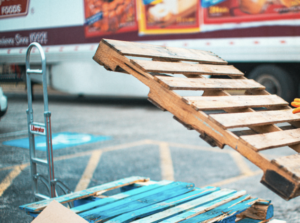 A person wearing gloves and a mask lifts a wooden pallet onto a stack of pallets in an outdoor parking lot near a truck, pondering, "How Much Does It Cost to Get Custom Pallets Made in Surrey?.