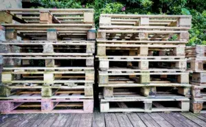 Stacks of custom pallets are piled outdoors on a wooden surface, with green foliage in the background.