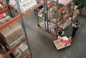 Two workers in a warehouse move a pallet loaded with paint buckets, surrounded by shelves stacked with boxes and supplies—all supported by heat treated custom pallets for added safety and durability.