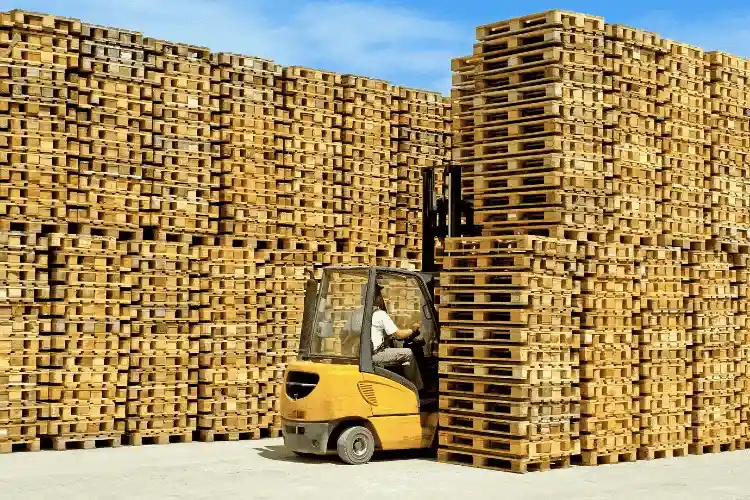 A forklift moves wooden pallets in an outdoor storage area at a Pallet Company in Surrey, with large stacks of pallets under a clear sky.