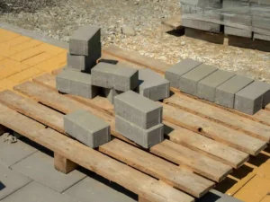 Gray concrete bricks are stacked in two uneven piles on a wooden pallet outdoors, showcasing custom pallet construction, with some bricks scattered and more construction materials visible in the background.