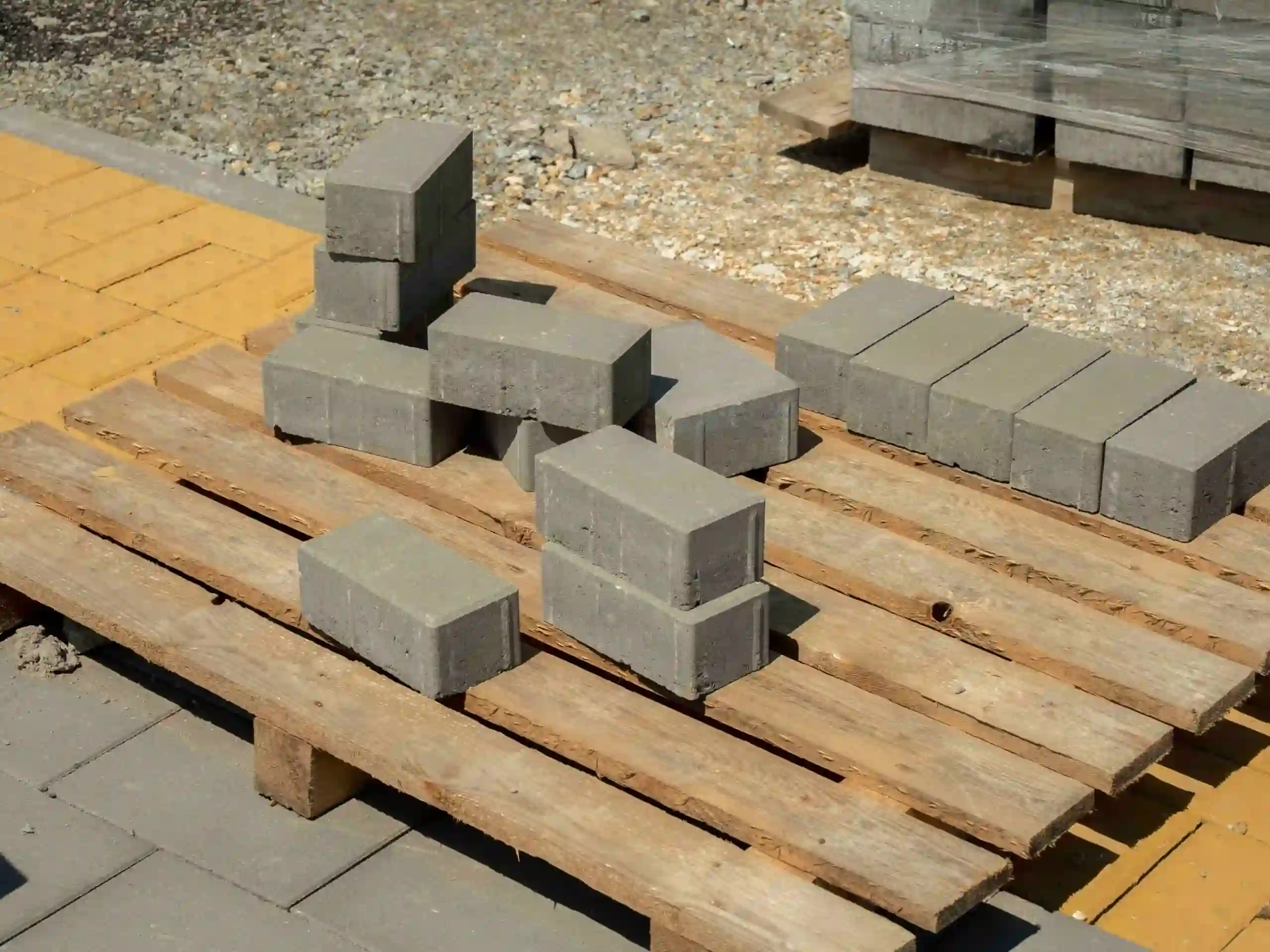 Gray concrete bricks are stacked in two uneven piles on a wooden pallet outdoors, showcasing custom pallet construction, with some bricks scattered and more construction materials visible in the background.