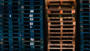 Stacked blue plastic pallets with barcode labels are on the left, and stacked natural wood pallets without labels are on the right, both seen from the side, illustrating a range of custom pallet options.
