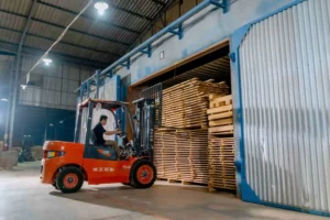 A person operates a red forklift to move stacked wooden planks, including Order Custom Pallets, into a large warehouse with open blue and white doors.