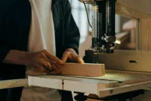 A person uses a band saw to cut a piece of wood in a workshop, skillfully working on repairs and creating custom pallets.
