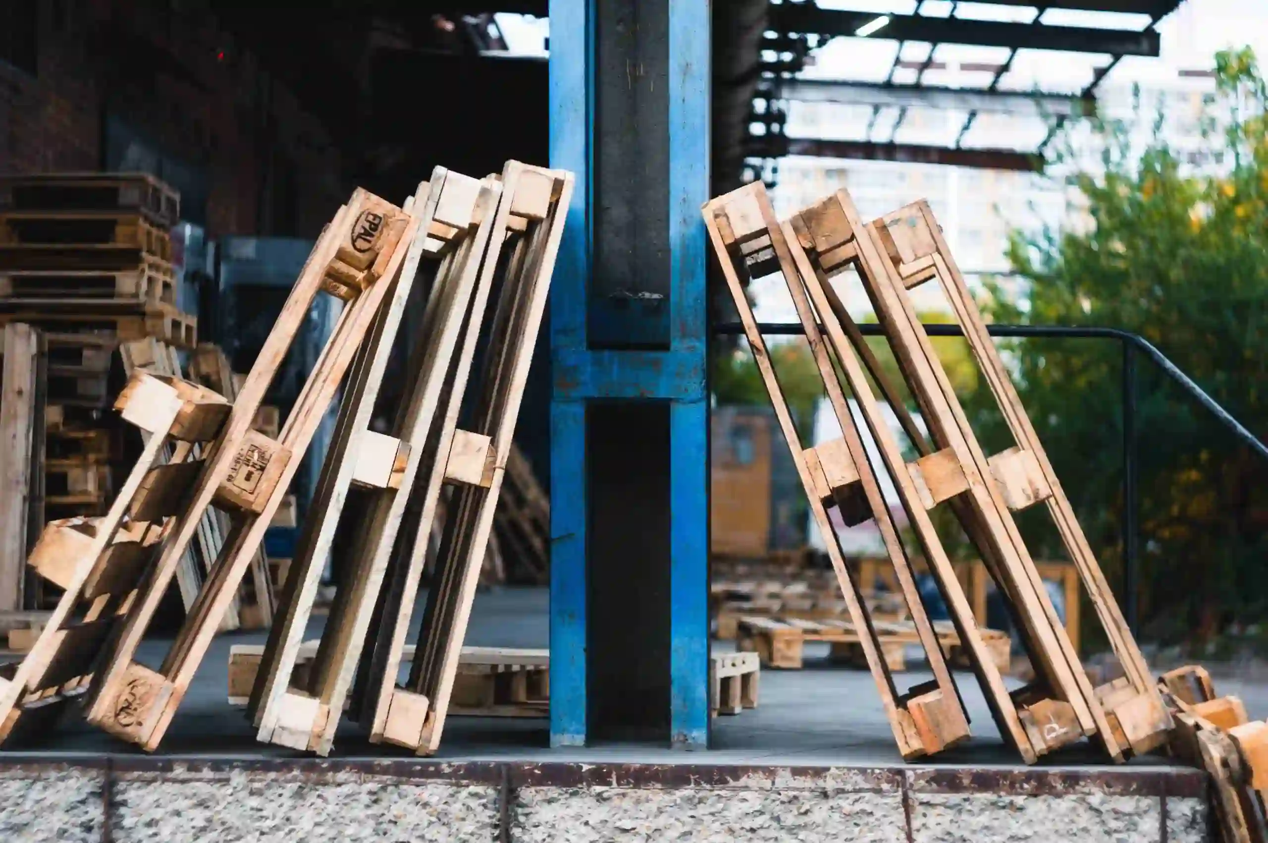 Two stacks of wooden pallets lean against a blue metal post in an industrial outdoor setting, hinting at the common problems with custom pallets, such as durability and proper storage solutions.