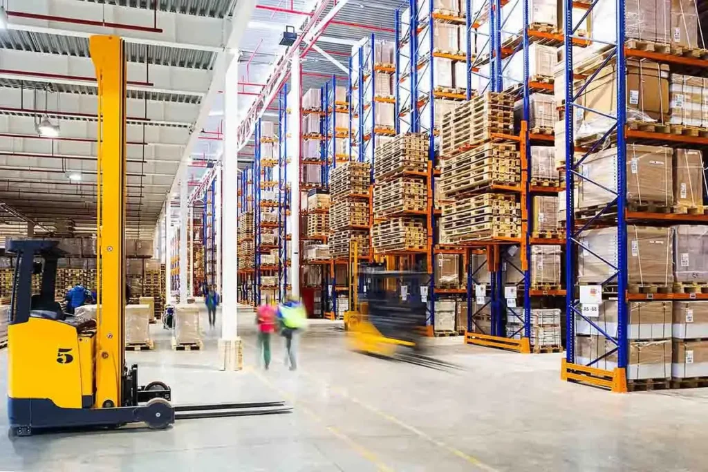 Warehouse interior with tall racks holding pallets and boxes—many supplied by a leading Pallet Company in Surrey—yellow forklifts operating, and a few workers in high-visibility vests moving through the space.