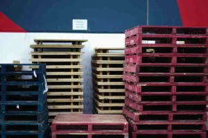 Stacks of wooden pallets, some painted red and blue, are arranged against a wall with a sign that reads "BROKEN PALLETS THIS SIDE," next to an area where you can store custom pallets for easy organization.