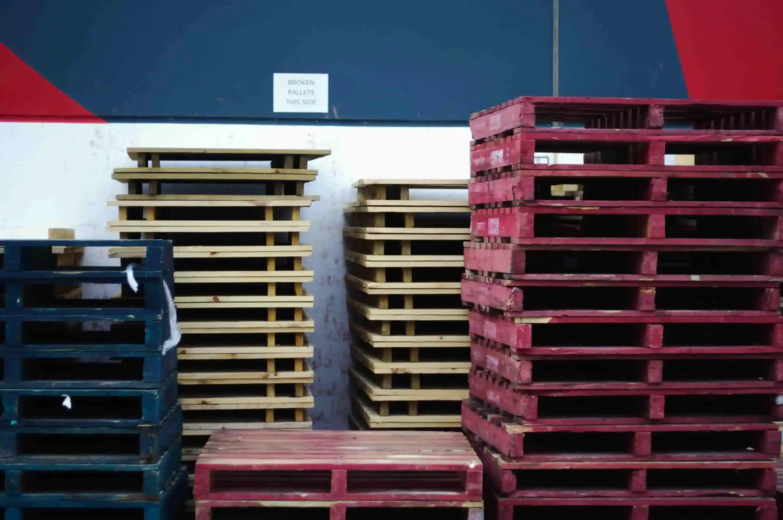 Stacks of wooden pallets, some painted red and blue, are arranged against a wall with a sign that reads "BROKEN PALLETS THIS SIDE," next to an area where you can store custom pallets for easy organization.