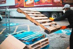 A person wearing gloves and a mask stacks a custom pallet onto others in a parking lot near a truck, carefully observing weight limits.