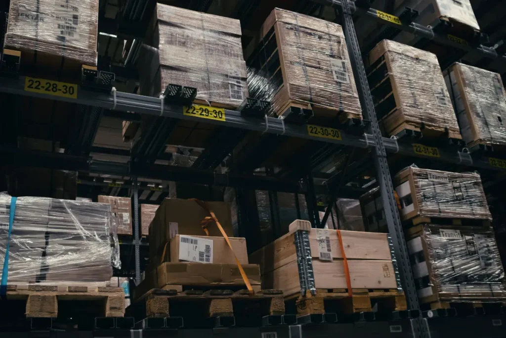 Wooden crates and cardboard boxes wrapped in plastic are stacked on shelves in a warehouse with numbered yellow labels, efficiently organized by a leading pallet company in Surrey.