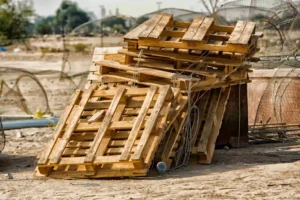 A stack of wooden pallets, including some old damaged custom pallets ready to dispose, is piled on sandy ground outdoors, with metal wires and debris scattered nearby.
