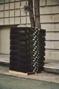 A stack of black heat-treated custom pallets sits on a sidewalk near a tree, in front of a beige stone building with a closed door.