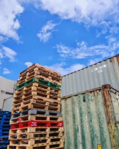 Tall stacks of wooden pallets—part of our custom pallet solutions—are placed beside green and gray shipping containers under a partly cloudy sky.