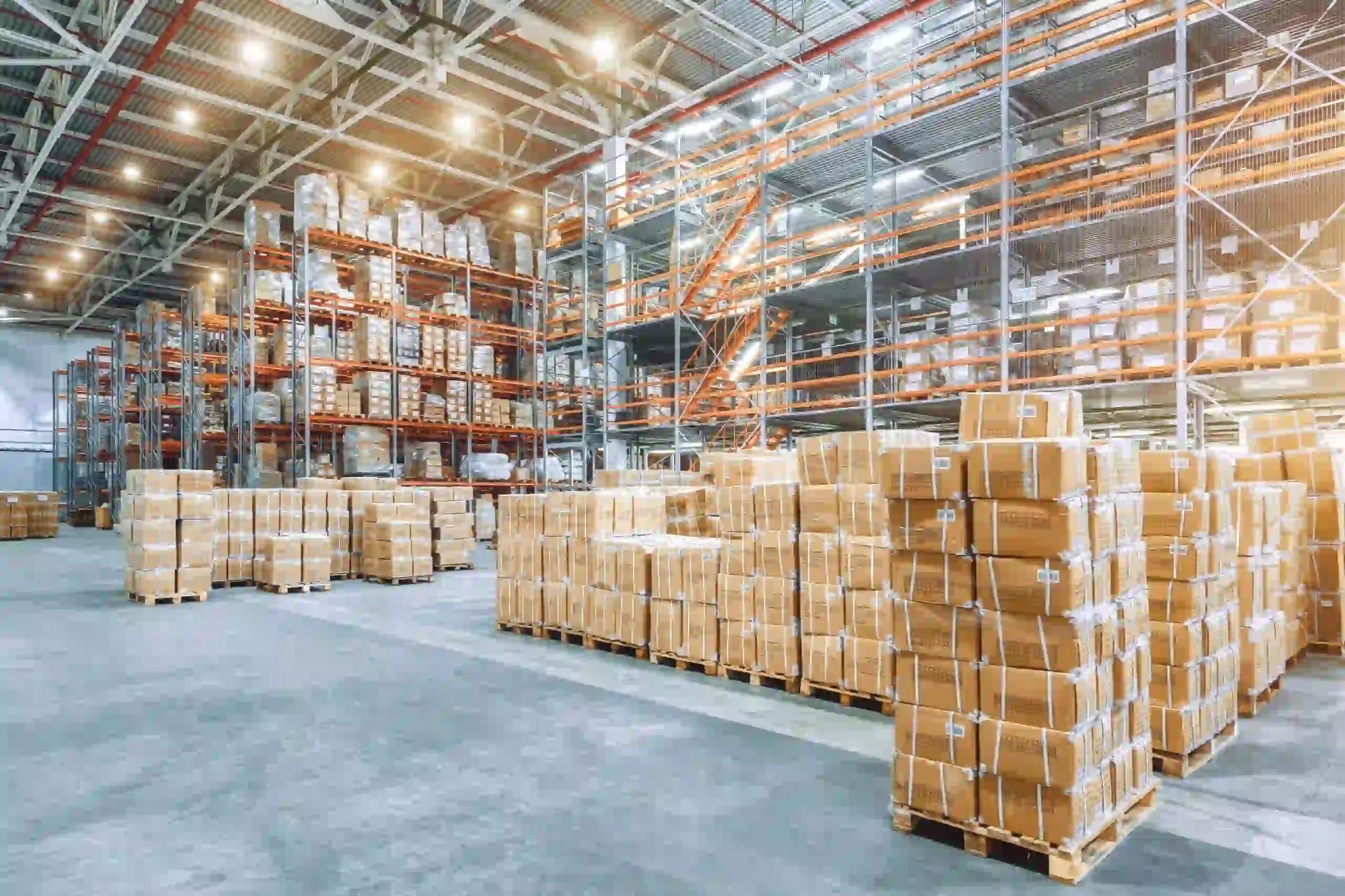 Large warehouse interior with tall metal racks filled with boxes and pallets from a leading Pallet Company in Surrey; additional stacked cardboard boxes are visible on the concrete floor. Bright overhead lighting illuminates the space.