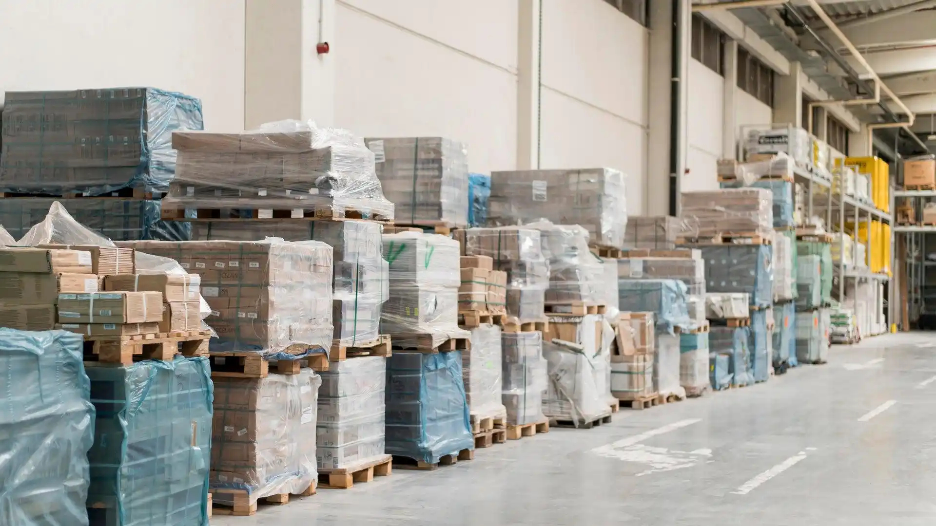 Stacks of goods wrapped in plastic sit on wooden pallets along the wall of a spacious warehouse with high ceilings and concrete floors, maintained by a leading pallet company in Surrey.