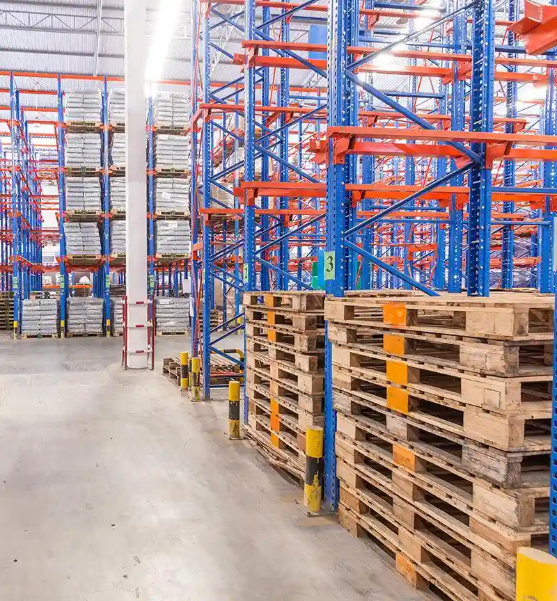 Warehouse interior by a leading Pallet Company in Surrey, featuring blue and orange racking, stacked pallets on the right, and shelves filled with goods in the background. Concrete floor and bright overhead lighting complete the organized storage space.