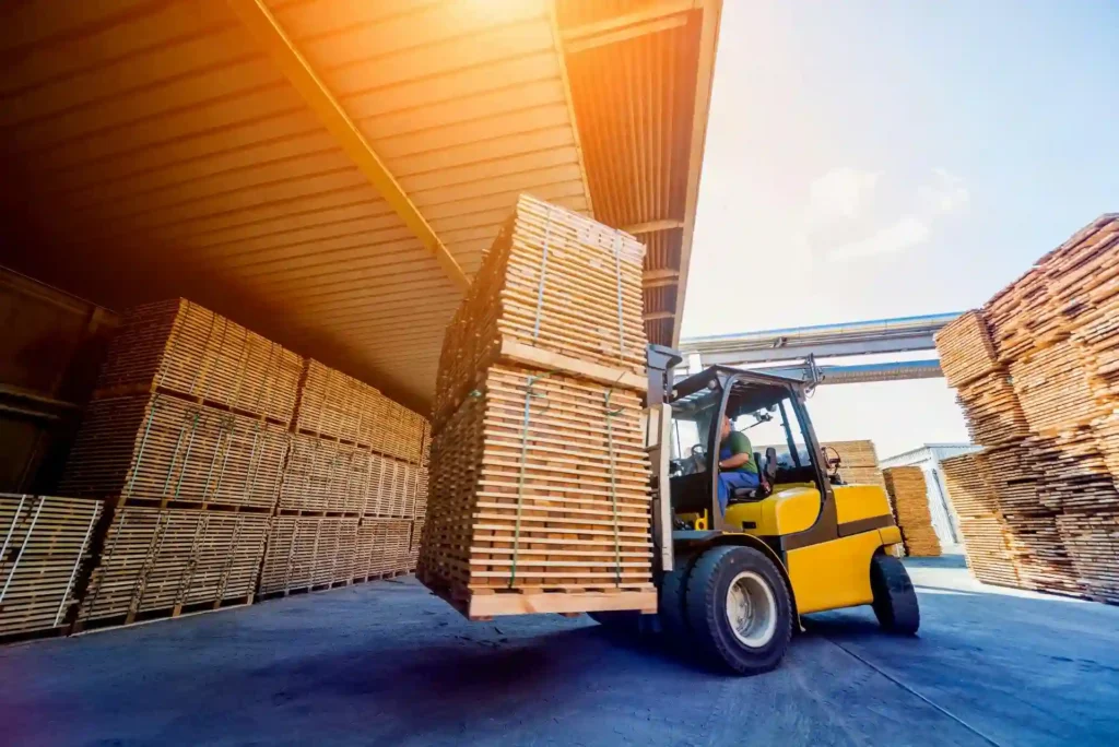 A forklift carries a large stack of wooden boards outside a warehouse, with sunlight shining through the roof—another busy day at a leading pallet company in Surrey.