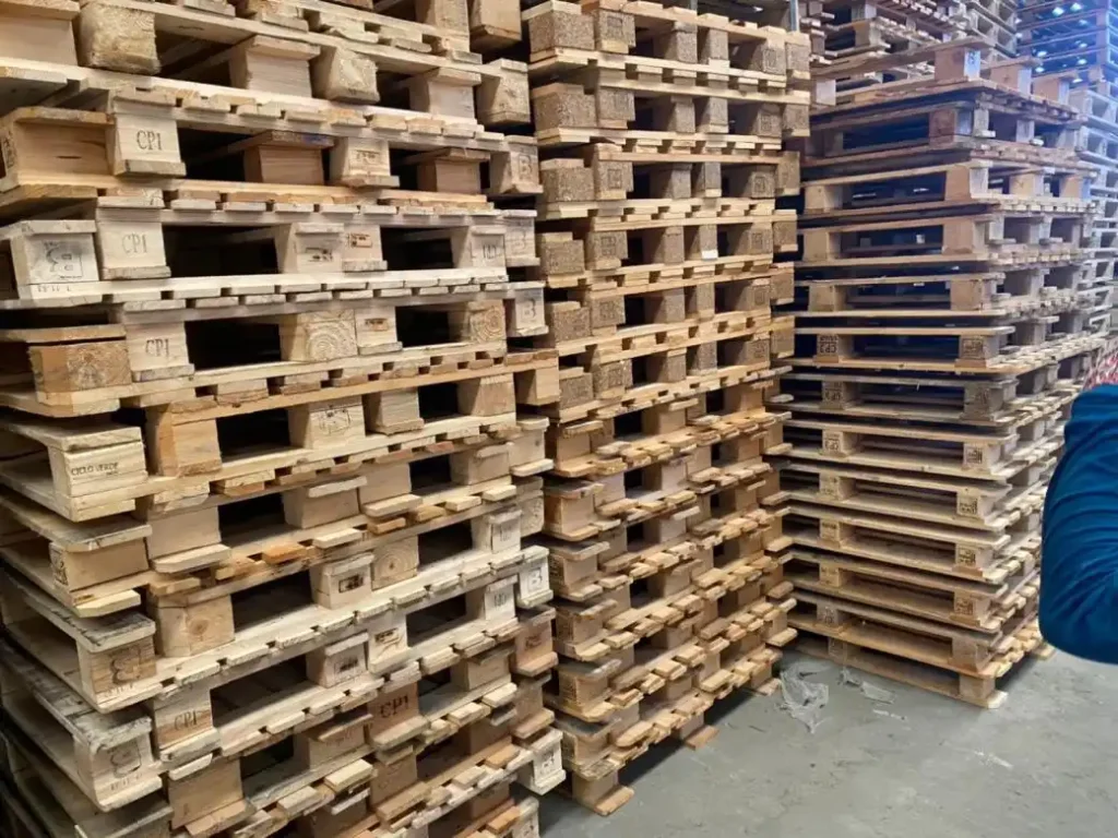 Stacks of wooden pallets are piled high against a wall on a warehouse floor, with some stamping visible on the sides of the pallets—evidence of their quality sourcing from a leading Pallet Company in Surrey.