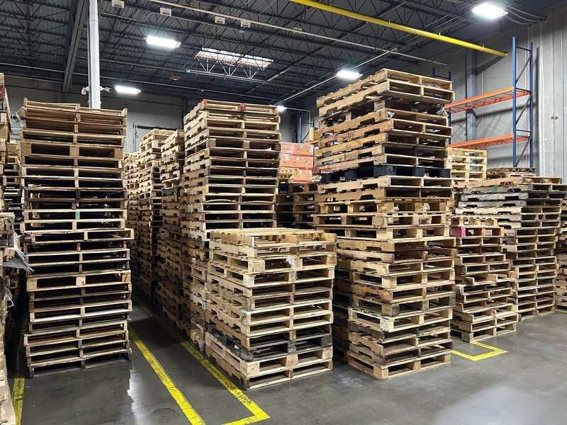 Stacks of wooden pallets are arranged in rows inside a warehouse with high ceilings and concrete floors, showcasing the impressive inventory managed by a leading pallet company in Surrey.