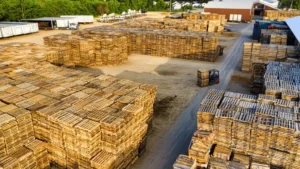 Aerial view of a storage yard with large stacks of wooden pallets, several trucks parked along the edge, and a forklift moving pallets on a dirt road.